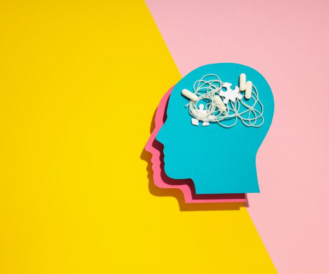 A cut-out of a blue head on a yellow and pink background. Inside the head are white puzzle pieces, pills and string.