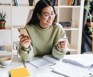 A woman sitting at a desk, wearing a mint green sweater. She is holding a credit card in one hand and a cell phone in the other hand, smiling.