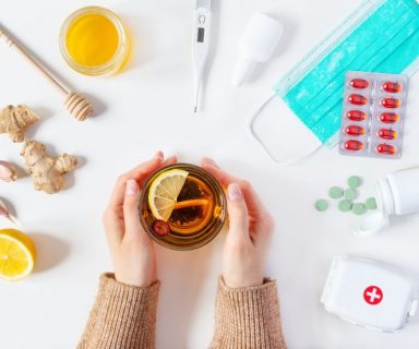 Two hands holding a mug of tea, with natural and medical remedies for cold flu treatment on the white table around them. There are different pills, ginger, lemons, a face mask, and a thermometer.