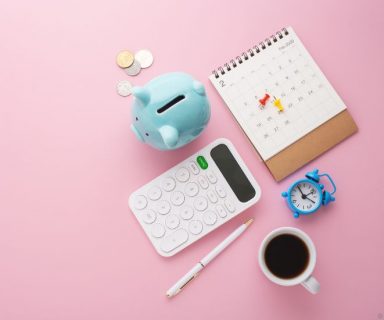 A blue piggy bank, calendar, calculator, pen, and mug of coffee on a pink background.