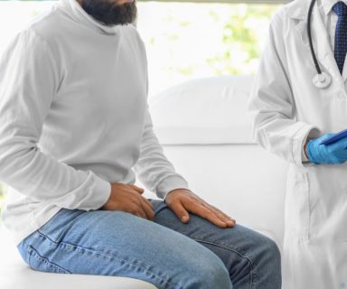 A man sitting on a table, fully stressed, with a hand over his groin. A doctor is standing beside him.