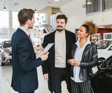 A couple talking to a car salesman inside a car dealership.
