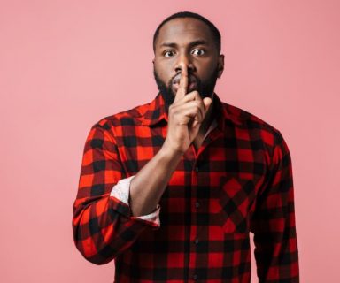 A man in a red and black plaid shirt with a finger up to his mouth, like he has a secret.